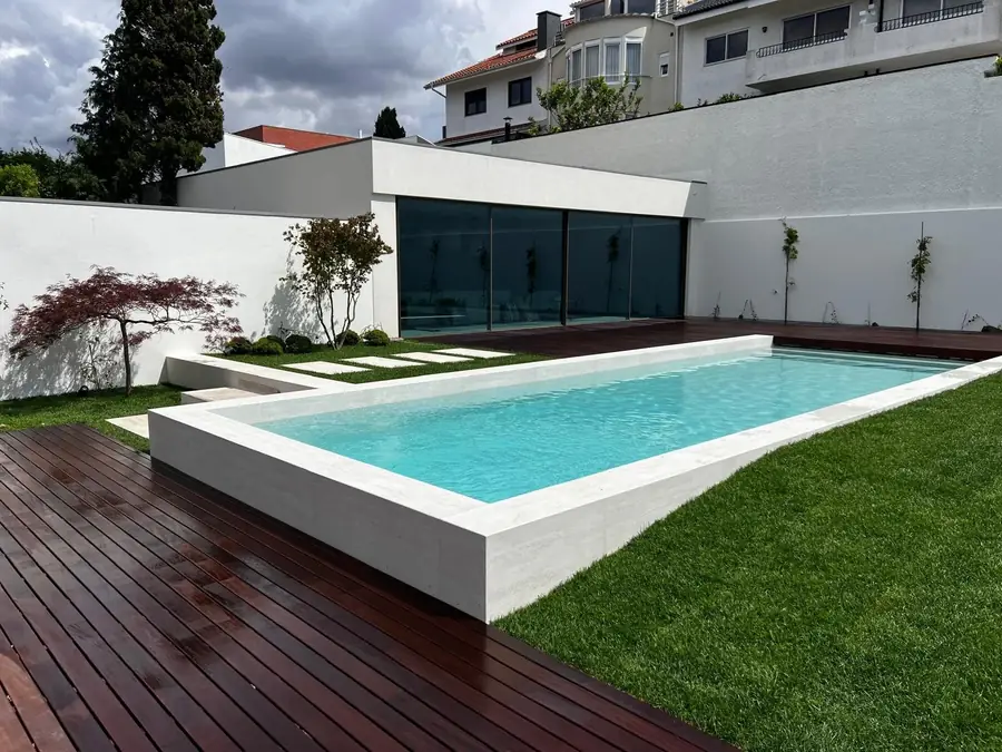 A rectangular pool edged in white stone, set into a lawn beside a hardwood deck and a white house.